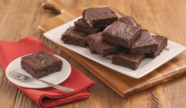 Fotografia em tons de marrom e vermelho de uma bancada de madeira com uma tábua de madeira, sobre ele um papel manteiga com pedaços de brownie. Ao  fundo um prato com um pedaço de brownie e um garfo.