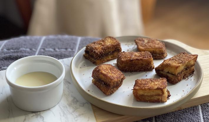 Foto aproximada de um prato com pedaços de Pão com Canela French Toast, ao lado de um potinho pequeno com um pouco de Leite Moça, tudo sobre uma toalha cinza quadriculada