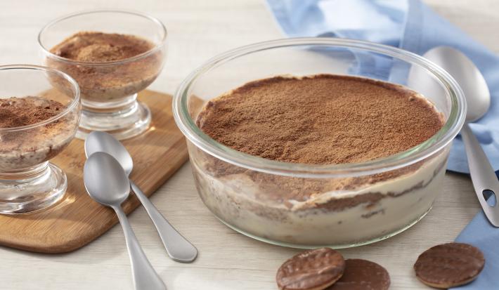 Fotografia em tons de azul em uma bancada de madeira clara, um paninho azul, uma tábua de madeira, um recipiente de vidro redondo com o tiramisú feito com calipso. Ao lado, em cima da tábua, duas taças com o tiramisú entro.