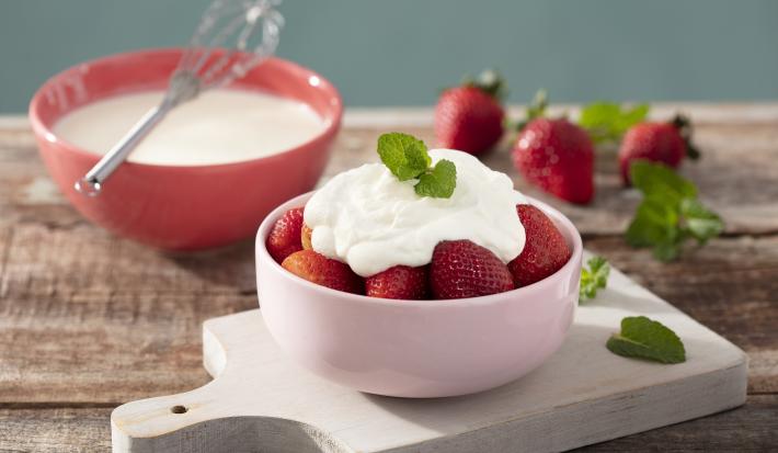 Fotografia em tons de vermelho em uma bancada de madeira clara, uma tábua de madeira branca, um potinho rosa claro com morangos dentro cobertos com creme de leite fresco. Ao fundo, um potinho vermelho com o creme de leite fresco dentro dele.