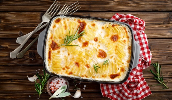 Em cima de uma mesa, refratário com batatas com queijo gratinado com molho branco, decorado com alecrim.