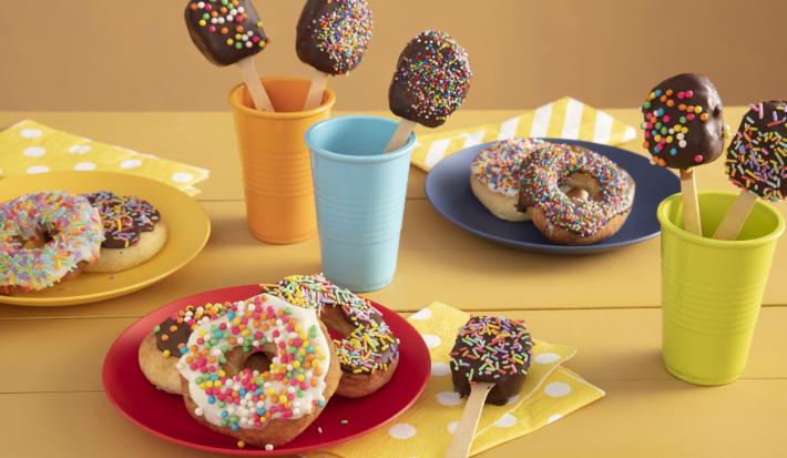 Fotografia em tons de amarelo em uma bancada de madeira amarela com três copos coloridos, azul, laranja e verde com pirulitos de chocolate dentro dele. Ao centro, um prato vermelho com três donuts cobertos com granulados coloridos.