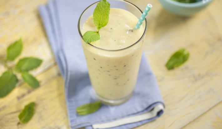 Fotografia em tons de marrom e azul de uma bancada de madeira com um paninho azul, sobre ele um copo com smoothie com um canudo. Ao lado folhinhas de hortelã e um recipiente redondo com hortelã.