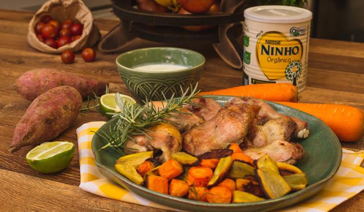 Fotografia em tons de marrom e verde de uma bancada de madeira, paninho xadrez amarelo e branco e um prato verde com pedaços de frango, cenoura e batata-doce. Ao fundo lata de NINHO e potinho com molho.