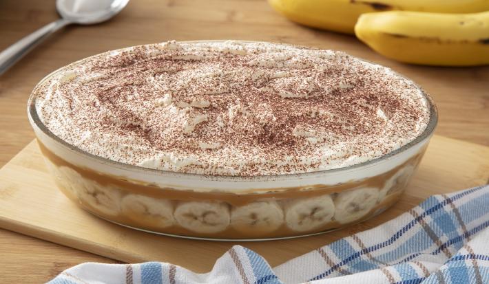 Foto da receita de pavê de banoffee servida em uma travessa de vidro grande sobre uma tábua de madeira em cima de uma mesa de madeira com um paninho quadriculado ao lado