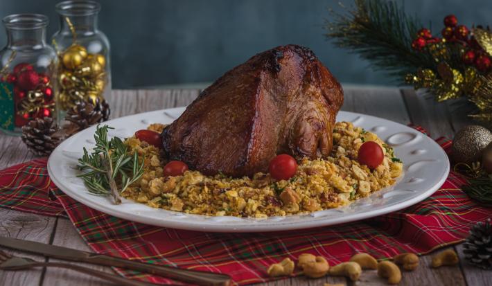 Foto da receita de pernil com farofa crocante, servido em um prato branco redondo, sobre uma mesa de madeira toda decorada com itens natalinos. Há algumas castanhas de caju espalhadas na mesa e um conjunto de talheres à frente do prato