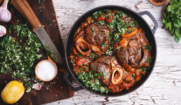 Fotografia vista de cima uma panela de ferro com ossobucos com molho e salsinha por cima. Ao lado, uma tábua de madeira em tom escuro, e em cima, uma faca, salsinha picada, duas cabeças de alho, sal grosso e um limão siciliano sem a casca.