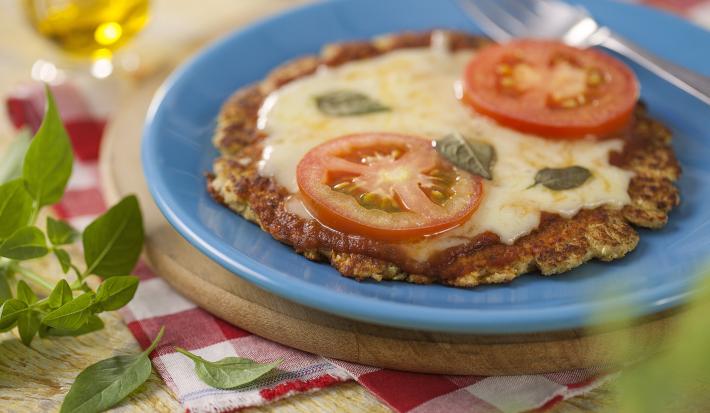 fotografia em tons de azul, vermelho e marrom de uma bancada marrom vista de cima, contém uma tábua redonda, um prato redondo azul com uma mini pizza coberta com queijo, duas fatias de tomate e manjericão e ao lado um garfo para servir.