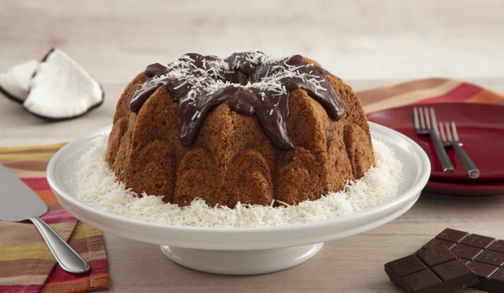 Fotografia em tons de vermelho em uma bancada de madeira clara com um paninho listrado colorido ao lado, uma espátula para bolo apoiada nele. Ao centro, um suporte para bolo branco com o bolo de coco com cobertura de chocolate em cima dele.