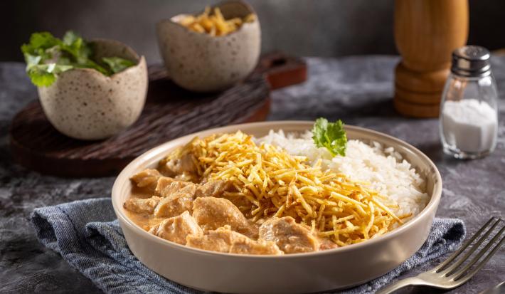 Imagem de um prato redondo com arroz, batata palha e strogonoff de frango cremoso. Compondo a imagem temos um saleiro, um potinho de salsinha e um potinho de batata palha.
