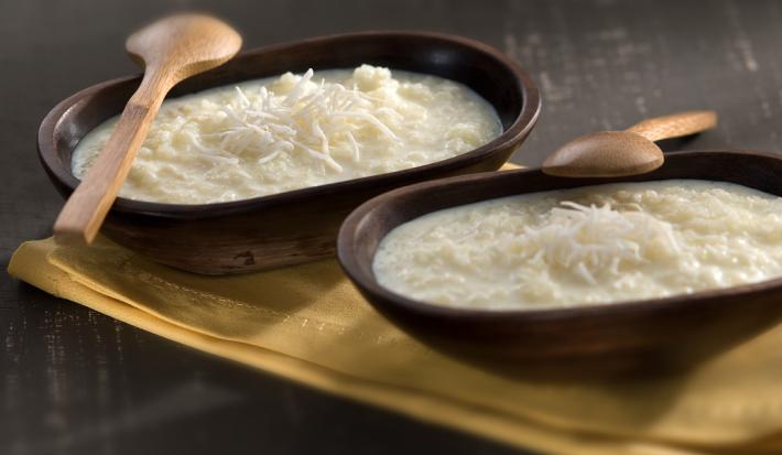 Fotografia em tons de marrom, branco e bege, com dois recipientes de madeira escura com tapioca, coco ralado e duas colheres de pau sobre guardanapo bege, tudo sobre bancada em tons de cinza.