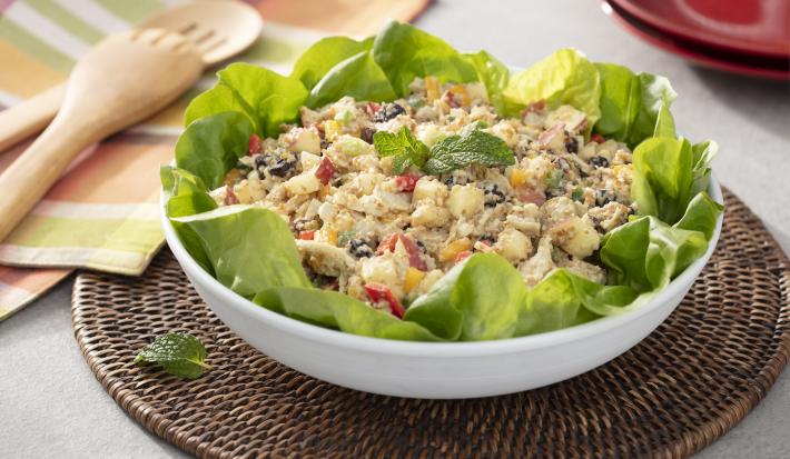 Fotografia em tons de verde em uma bancada de madeira, um pano com listras coloridas, um jogo americano redondo de vime escuro e um recipiente branco redondo fundo com a salada marroquina com vários legumes dentro dela.