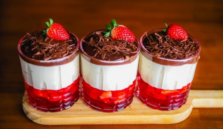 Fotografia em tons de marrom com três potes de vidro ao centro. Dentro de cada pote existe uma sobremesa em camadas feita com gelatina de morango, creme branco e coberta com ganache de chocolate.