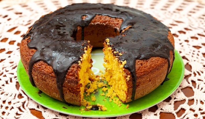 Bolo redondo coberto com uma rica cobertura de chocolate, colocado em um prato verde vibrante. O bolo tem um aspecto rústico e é apresentado sobre uma toalha de mesa de renda branca.