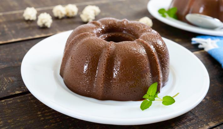 Fotografia de um pudim pequeno de chocolate meio amargo e folhas de hortelã que estão em cima de um prato raso de vidro na cor branco. Ao fundo, o mesmo pudim sobre um prato, um pano azul e algumas pipocas sobre uma mesa de madeira de tom escuro.
