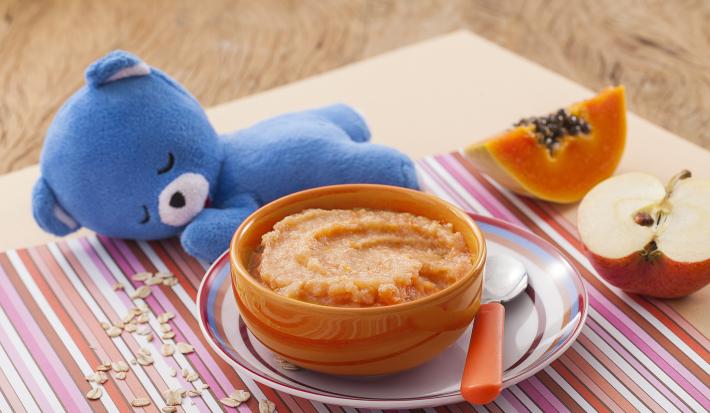 Fotografia em tons de laranja e azul de uma bancada de madeira com um paninho listrado colorido, um potinho laranja com o purêzinho de mamão, atrás um ursinho de pelúcia azul. Ao fundo, meio pedaço de maçã e meio pedaço de mamão.