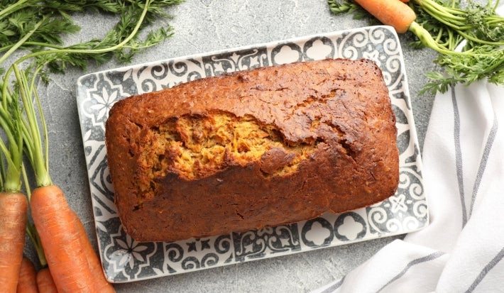 Bolo de cenoura retangular, dourado e rústico, com superfície rachada e pedaços de cenoura visíveis. Está sobre prato preto e branco, cercado por cenouras frescas e ao lado de um pano branco com listras cinza.