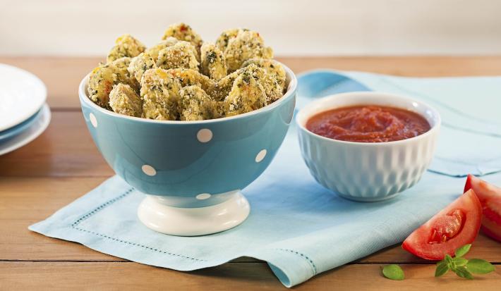 Fotografia em tons de marrom e azul claro de uma mesa de madeira vista de frente, contém um pano cor azul claro e por cima um recipiente redondo cor azul com bolinhas brancas com pedaços de nuggets, ao lado um pote branco com molho de tomate.