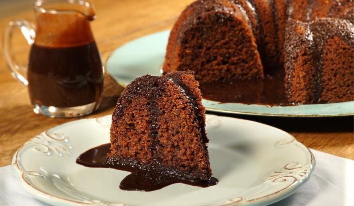 Fotografia em tons de marrom, branco e azul de uma bancada de madeira com um paninho branco, sobre ele um prato redondo branco com uma fatia de bolo. Ao fundo um prato redondo azul com o bolo. Ao lado uma jarra com calda de chocolate.