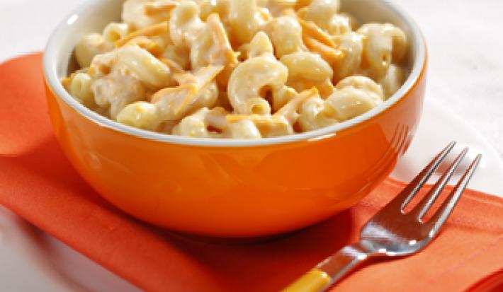 Fotografia em tons de laranja e branco, um prato redondo branco com um paninho laranja dobrado em cima, um garfo com cabo amarelo e um potinho laranja com o macarrão cremoso dentro.