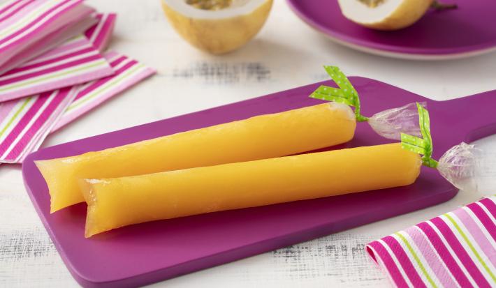 Fotografia em tons de branco e roxo de uma bancada de madeira branca e uma tábua roxa, sobre ela dois geladinhos de maracujá. Ao lado paninhos listrados em tons de roxo e branco, ao fundo um prato redondo roxo com um maracujá cortado ao meio.