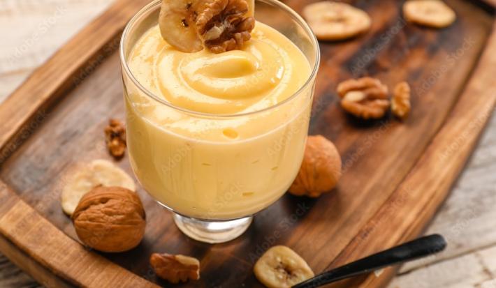 Fotografia de uma flan de nozes e leite MOÇA com uma rodela de banana e uma noz por cima dentro de um recipiente de vidro decorativo. O creme está sobre uma tábua de madeira com várias nozes espalhadas por cima, juntamente com uma colher de sobremesa.