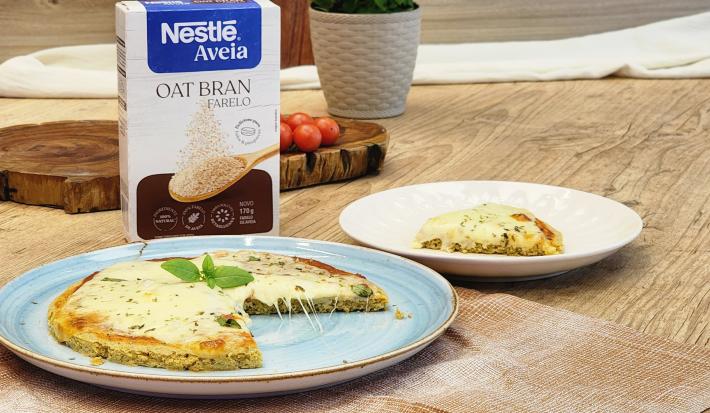 Fotografia em tons de marrom com um prato azul ao centro. Em cima do prato existe uma pizza com massa de Aveia coberta com queijo muçarela e orégano. Ao fundo existe uma embalagem de Farelo de Aveia NESTLÉ.