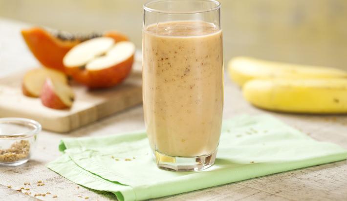 Fotografia em tons de rosa, verde e branco, com copo ao centro com vitamina sobre guardanapo verde, no entorno duas bananas, maçã cortada em pedaços e um pedaço de mamão sobre peça de madeira, potinho com linhaça, tudo sobre bancada em tons de branco.