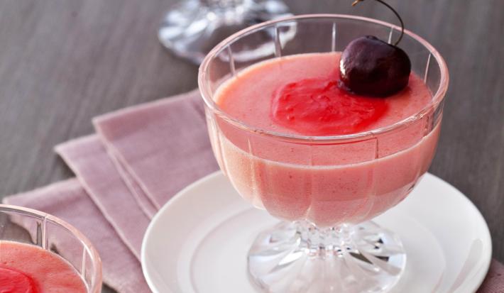 Fotografia em tons de vermelho em uma mesa de madeira com um pano rosa escuro e um prato pequeno branco apoiado nele. Em cima do prato, uma taça de vidro com a mousse de cereja e uma cereja por cima para enfeita. Ao lado e ao fundo, mais duas taças.