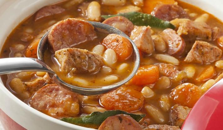 Fotografia em tons de branco e marrom de um recipiente redondo branco com cassoulet de feijão branco, sobre ele uma concha com uma porção do feijão.