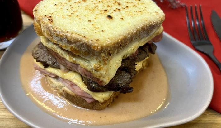 Foto da receita de Francesinha. Oberva0se um prato de cerâmica cinza com o sanduíche por cima, com queijo gratinado e um molho amarronzado.
