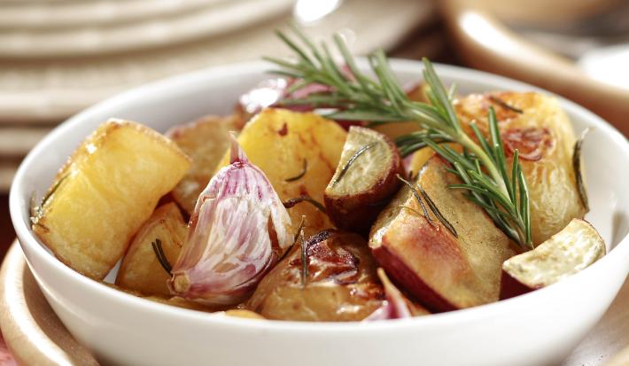 Fotografia em tons de bege em uma bancada de madeira escura, uma toalha vinho, um pote bege e um branco redondo em cima com o mix de batatas assadas dentro dele.