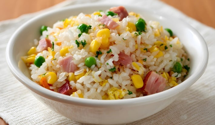 Em cima de uma mesa, tigela redonda com porção de arroz com presunto cortado, ervilha, cenoura picada e milho.