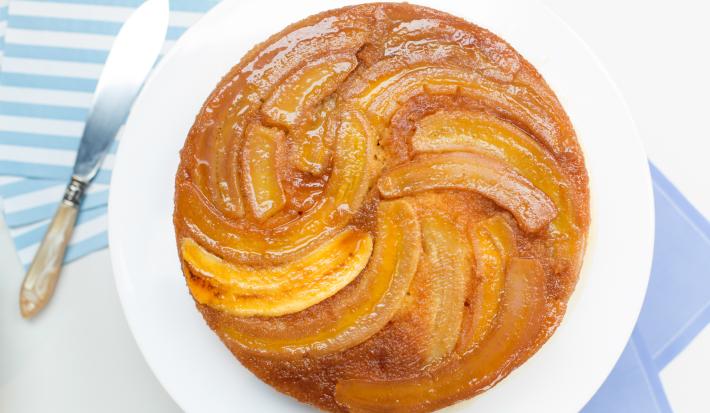 Fotografia em tons de azul e branco de uma bancada branca com um paninho azul, sobre ela um prato redondo branco com um bolo de banana. Ao lado um paninho listrado e uma faca.