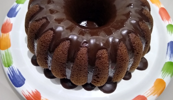 Foto em tons de marrom da receita de bolo de café servida com uma cobertura de chocolate por cima e sobre um prato branco com a borda colorida
