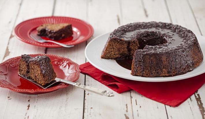 Fotografia em tons de vermelho em uma bancada de madeira clara com um prato branco ao centro com o bolo de chocolate cortado ao meio e um paninho vermelho embaixo. Ao lado, dois pratos vermelhos com uma fatia de bolo em cada um.