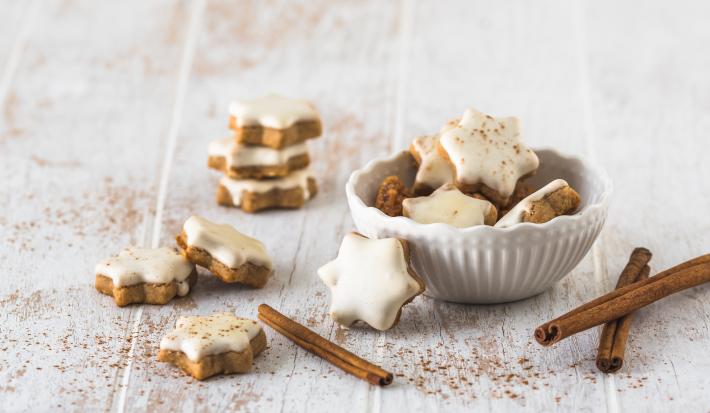 Fotografia de várias unidades de biscoito alemão em formato de estrela e uma pasta branca por cima. Uma parte dos biscoitos estão dentro de um pote de vidro branco, e o resto está espalhado em volta do refratário, sobre uma mesa de madeira branca.