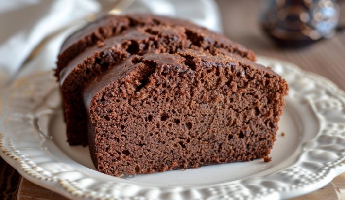Três fatias de bolo de chocolate fofinho em um prato cerâmico branco. O bolo é úmido e denso, com superfície levemente rachada. Ao fundo, uma xícara desfocada complementa a cena aconchegante.