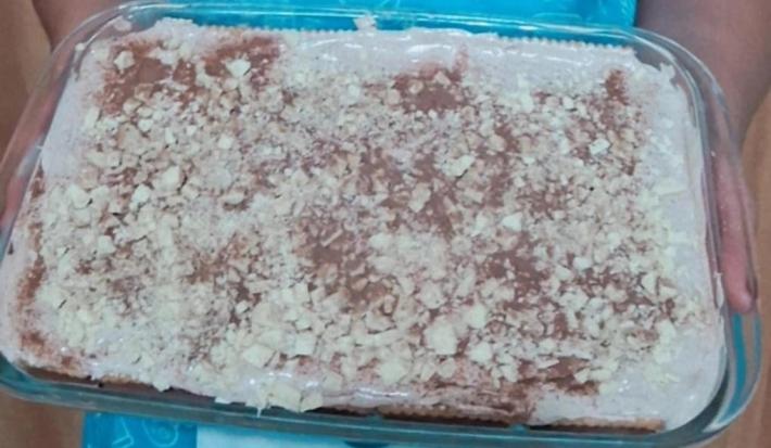 Fotografia de uma travessa de vidro retangular com camadas de sorvete e chocobiscuit. Por cima tem pedaços pequenos de chocolate branco e nescau polvilhado.