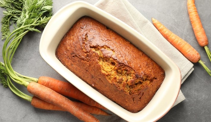 Bolo de cenoura retangular assado em uma forma de cerâmica branca. O bolo é dourado, com uma leve rachadura no topo, sugerindo uma textura macia por dentro. Ao redor da forma há cenouras frescas, inteiras com folhas verdes..