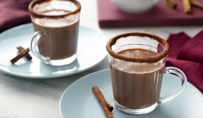 Fotografia em tons de marrom, em uma bancada de madeira de cor branca. Ao centro, um pires azul claro contendo uma xícara de chocolate quente com uma canela em pau ao lado. Ao fundo, um pano roxo e um pires com outra xícara.