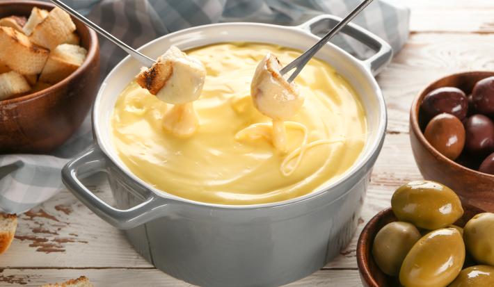 Fotografia em tons de branco com uma panela de fondue ao centro. Dentro da panela existe um fondue de queijo. Dois pedaços de pão estão sendo mergulhados no fondue de queijo. Ao fundo existe recipientes com azeitona e pedaços de pão.