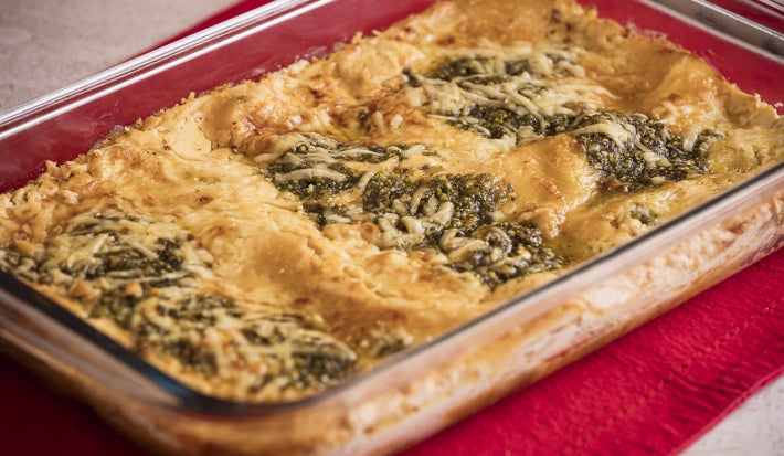 Foto da receita de Lasanha e Queijo Parmesão e Pesto, servida em uma travessa de vidro, sobre um pano vermelho.