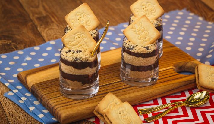 Imagem de uma bancada de madeira na qual há dois panos em tons azul claro e vermelho com texturas de bolinhas e listras. Há uma tábua pequena e sobre ela quatro copinhos com a receita em camadas de creme e biscoito triturado. Imagem de uma bancada de madeira na qual há dois panos em tons azul claro e vermelho com texturas de bolinhas e listras. Há uma tábua pequena e sobre ela quatro copinhos com a receita em camadas de creme e biscoito triturado.