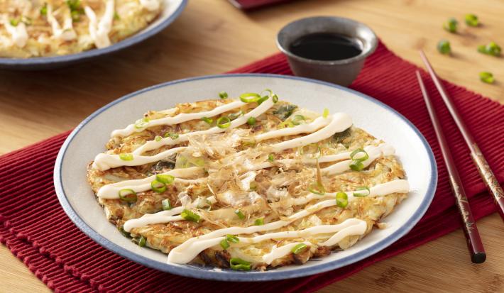 Fotografia em tons de vermelho em uma bancada de madeira com um jogo americano vermelho escuro, um prato branco com detalhes em azul ao centro com o okonomiyaki dentro dele e ao lado, um par de hashi. Ao fundo, um potinho com o molho de soja.