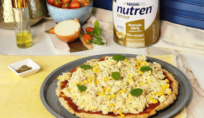 Fotografia em tons de amarelo com um recipiente circular ao centro. Dentro desse recipiente existe uma pizza de frango, queijo e manjericão. Ao fundo existe uma lata de Nutren Senior, cebolas e tomates