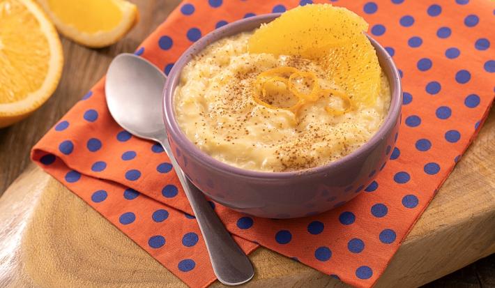 Foto em tons de laranja da receita de arroz-doce de laranja servida em um bowl de porcelana lilás sobre um pano laranja com bolinhas azuis com uma colher prateada ao lado. Ao fundo há laranjas cortadas sobre uma tábua de madeira