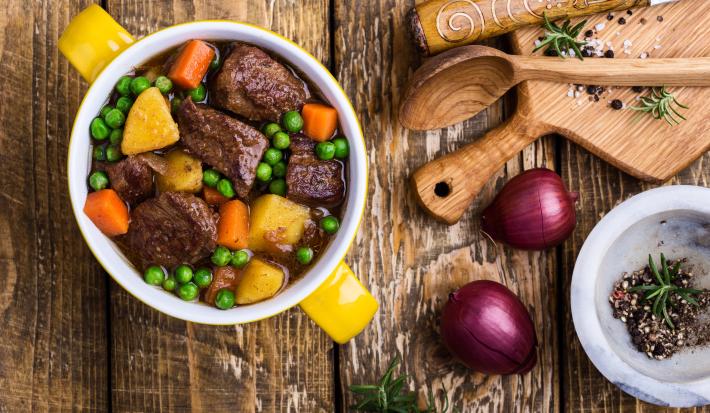 Fotografia vista de cima de um cozido de carne com ervilhas frescas, legumes e caldo de carne. Ao lado, duas cebolas roxas, uma tábua e colher de madeira e alguns temperos que estão dentro de um refratário fundo e pequeno, sobre uma mesa de madeira.