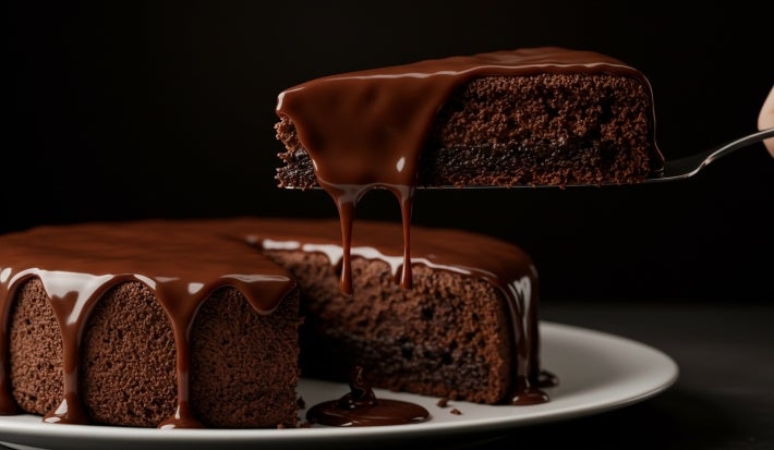 Um bolo de chocolate inteiro com cobertura de ganache brilhante. Um pedaço foi cortado e está sendo servido, revelando o interior macio e bem assado. A calda escorre de forma apetitosa pela fatia e pelas bordas do bolo.