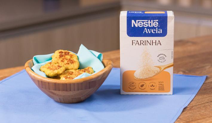 Fotografia em tons claros de uma mesa de madeira com um pano azul. Sobre o pano tem um recipiente de madeira fundo com um paninho azul claro com alguns nuggets saudáveis, ao lado da embalagem de NESTLÉ Farinha de Aveia.